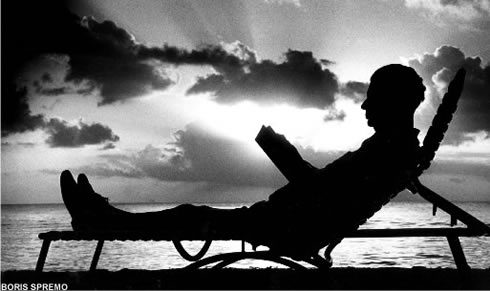 Silhouette of man on reclining chair with book at waterfront with sun going down