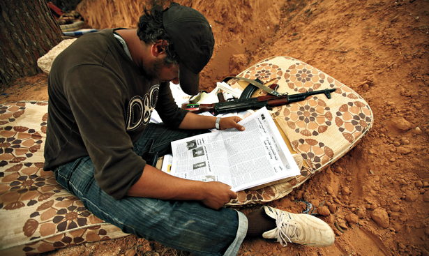 Man in dirt ditch on mattress next to gun reads newspaper
