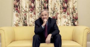 Man in suit on yellow cushion in front of floral curtains