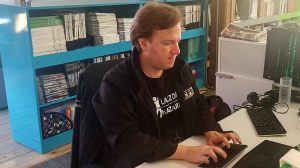 Joey Coleman sits at his desk. One of his computer monitors has a matrix background.