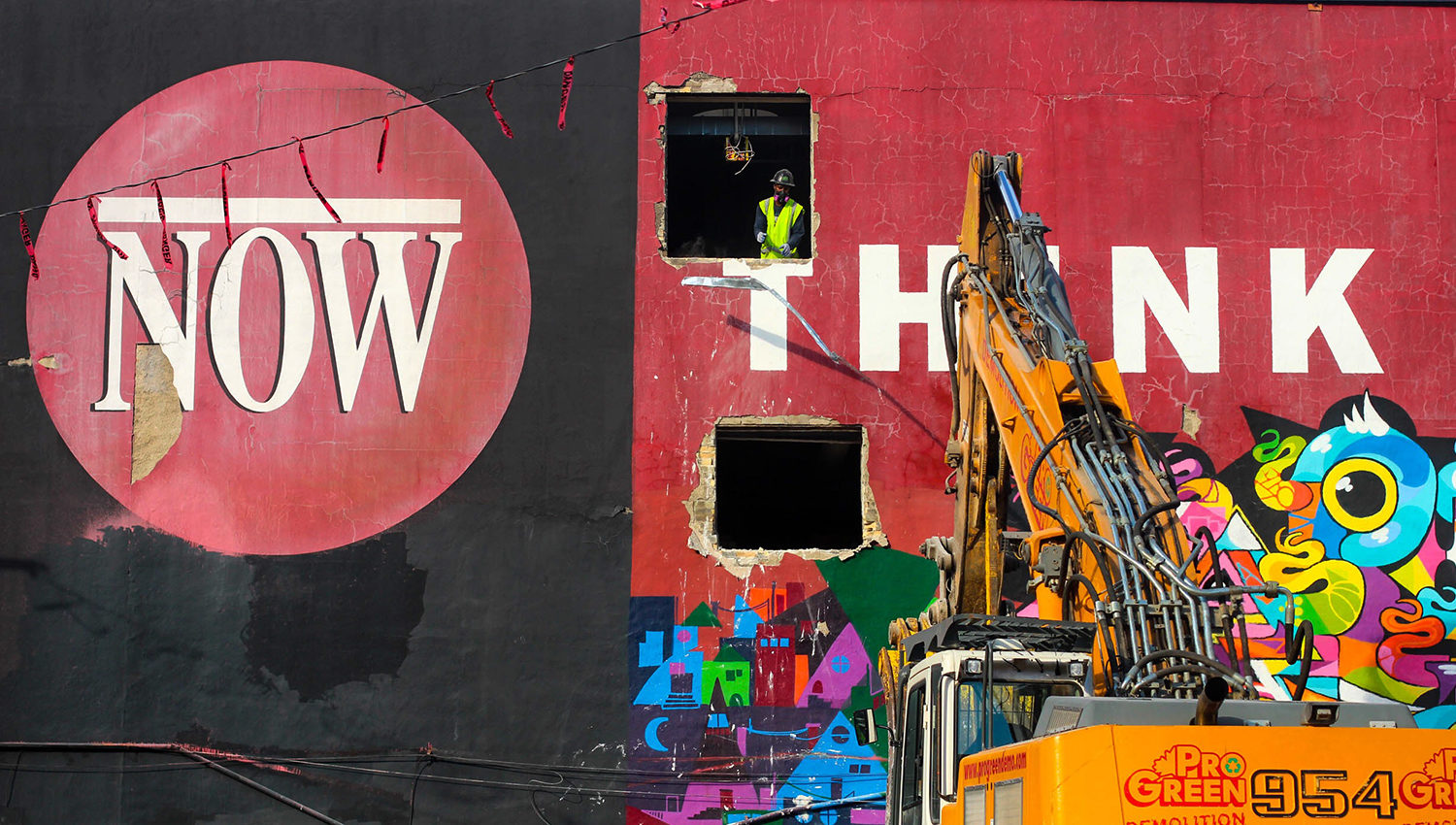 The Now red logo is presented on a black background on one half of the building. On the other side is the painted white word Think on a red background. A construction worker stands in an empty window frame. An excavator is positioned in front of the word Think.