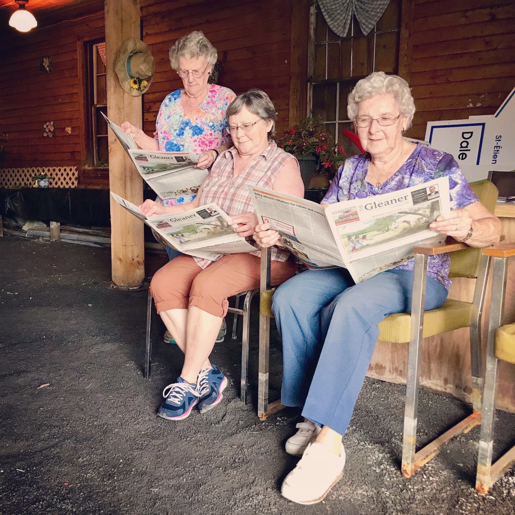 Three older white women, one standing and two sitting on comfy chairs, read copies of The Gleaner paper outside.