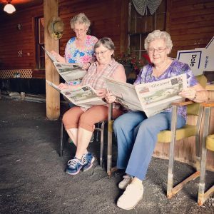 Three older white women, one standing and two sitting on comfy chairs, read copies of The Gleaner paper outside.