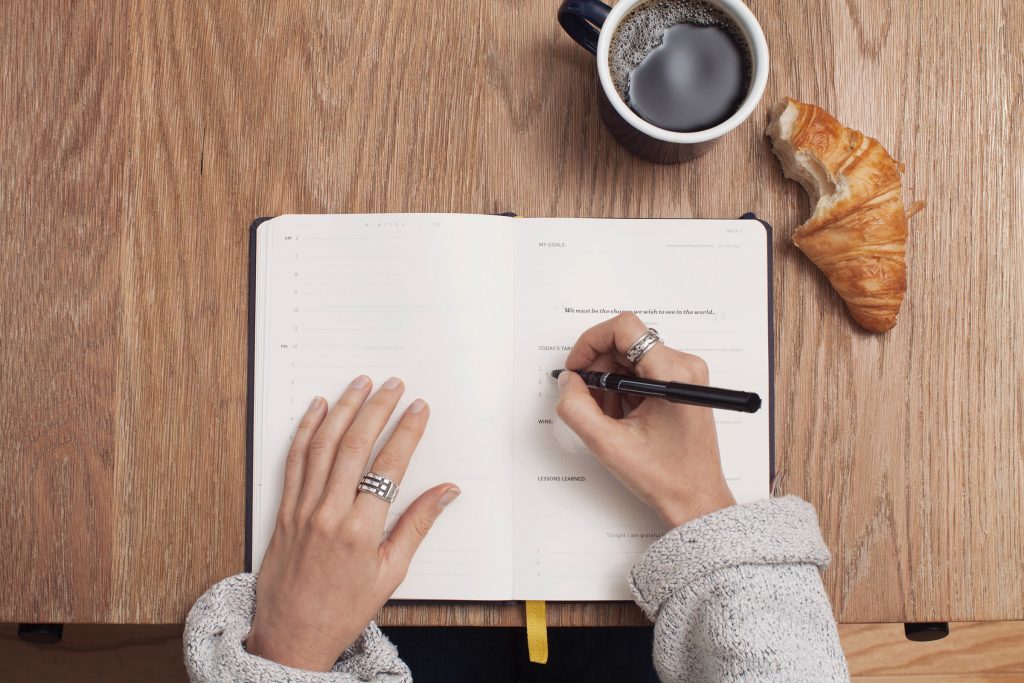 A pair of hands writes in an blank, open planner on a wooden table. There's a cup of coffee on the table as well.