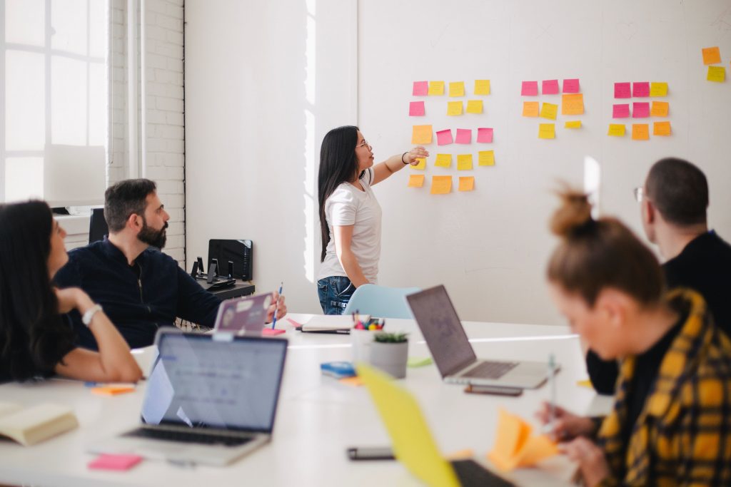 In a boardroom, multiple people sit around a white table. A short woman with dark hair stands at the front of the room pointing to coloured sticky notes on the wall.