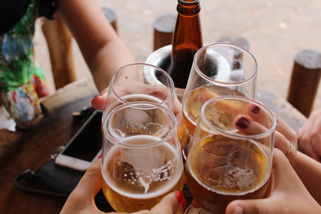 Four people doing cheers with four glasses of dark beer. There is a bottle of beer in the background.