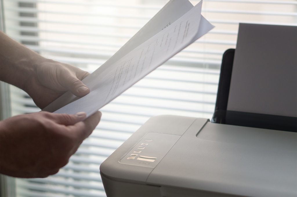 A person holds sheets of paper beside a printer.