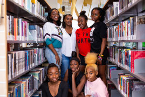 Annette Bazira-Okafor with 6 writers from Black Girls Magazine, all are standing between bookshelves and smiling at the camera