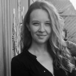 A black and white photo of a smiling woman with curly hair infront of a painted fence.